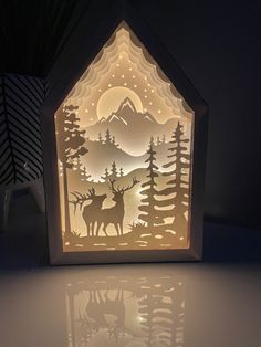 an illuminated paper house with deer and pine trees in the woods on it's side