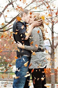 a man and woman kissing in front of trees with leaves falling from them on the ground