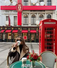 a woman sitting at a table drinking from a coffee cup in front of a red phone booth