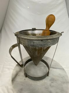 a metal strainer with a wooden spoon in it on a white surface, against a plain backdrop