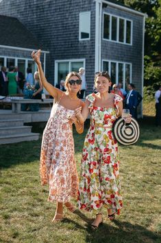 two women standing in front of a house with their arms up and one holding a straw hat