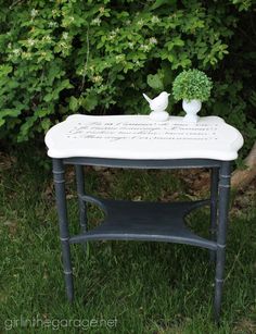 a small table with writing on it in front of some bushes and trees, near a tree