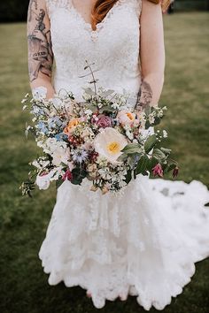 a woman in a wedding dress holding a bouquet of flowers and greenery with tattoos on her arm