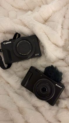 two cameras sitting on top of a white blanket next to each other and one is black