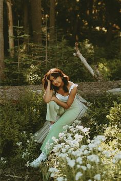 a woman sitting on a log in the middle of some flowers and trees with her hands behind her head