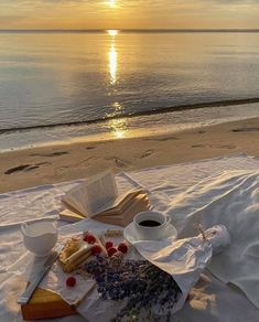 an open book and cup of coffee sit on a blanket at the beach as the sun sets