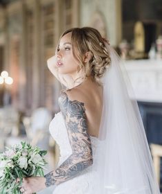 a woman in a wedding dress holding a bouquet and looking off to the side with tattoos on her arm