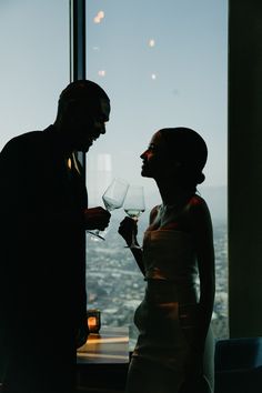 a man and woman standing next to each other holding wine glasses in front of a window