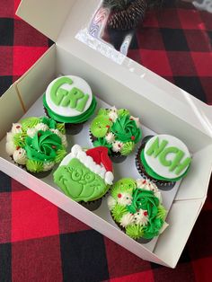 six decorated cupcakes in a white box on a red and black checkered tablecloth