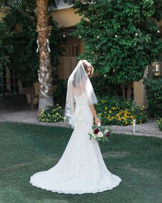 a woman in a wedding dress and veil standing on the grass with her back to the camera