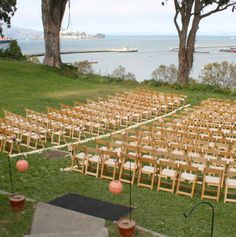 rows of wooden chairs are set up for an outdoor ceremony on the lawn near the water