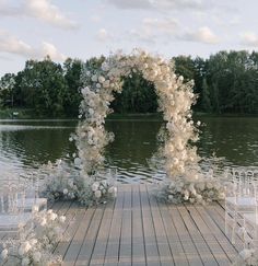 an outdoor wedding setup with white flowers and chairs on the dock by the water in front of some trees