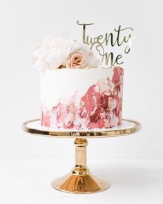 a white and pink cake with flowers on top sitting on a gold plate that says twenty one