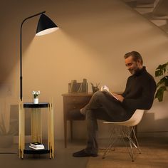 a man sitting in a chair reading a book next to a table with a lamp on it