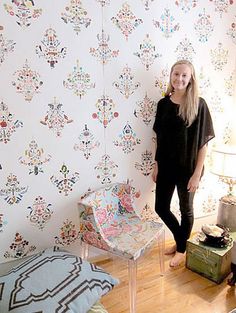 a woman standing in front of a wallpapered room with a chair and suitcase