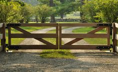 a wooden gate in the middle of a gravel road with trees and grass on either side