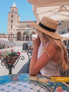 a woman sitting at an outdoor table with a hat on her head and flowers in the background