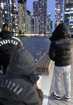 two people sitting on a bench looking at the water in front of some tall buildings