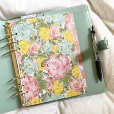 an open planner book with flowers on it and a pen next to it, sitting on top of a bed