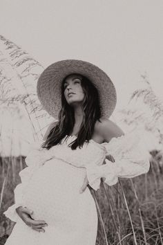 a pregnant woman wearing a hat and dress standing in tall grass with her hands on her hips