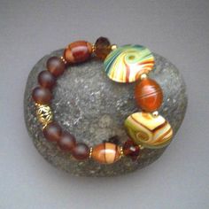 The beautiful 7-inch Southwestern-Boho-themed Mixed Stretch Bracelet of Porcelain Disks with vivid bands of Green and Dark Orange and Lampwork Glass beads is perfect for layering with other gold and gemstone bracelets.  An exquisite 16 X 12mm Handmade Orange with Gold-banded Lampwork Glass Bead is the central bead with Two large 25 X 20mm Polished Porcelain Disks with Green, Orange and Tan veins on either side.  Faceted 11 X 9 Czech Glass Rondel Beads in a Clear Sparkly Brown lead into 16 X 12mm Bohemian Round Brown Beads, Bohemian Czech Glass Polished Beads, Handmade Brown Czech Glass Beads, Bohemian Czech Glass Orange Beaded Bracelets, Bohemian Czech Glass Beads With Large Beads, Bohemian Czech Glass Orange Beaded Bracelet, Bohemian Orange Czech Glass Beaded Bracelets, Bohemian Orange Beaded Bracelets With Czech Glass, Bohemian Czech Glass Large Beads