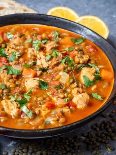 a black bowl filled with chicken and bean stew next to a slice of lemon wedges