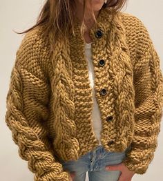 a woman standing in front of a white wall wearing a cardigan sweater and jeans