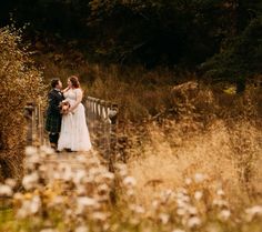 a bride and groom are standing on a bridge in the middle of some tall grass