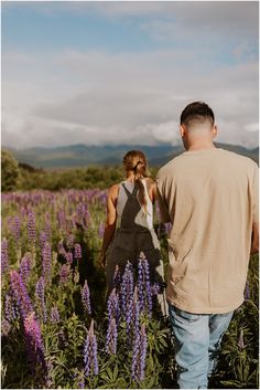 a man and woman walking through a field of purple flowers with mountains in the background