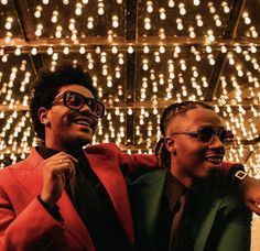 two men in suits and sunglasses standing next to each other with lights on the wall behind them