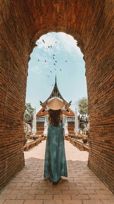 a woman in a blue dress and straw hat walking through an archway with birds flying overhead