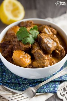 a close up of a bowl of food with meat and potatoes on a cloth next to a lemon wedge