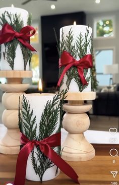 three white candles with red bows and pine branches on them, sitting on a table