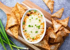 a bowl of dip surrounded by tortilla chips and chives on a cutting board