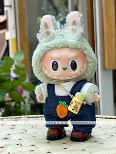 a small doll is sitting on a table wearing overalls and holding a carrot in her hand