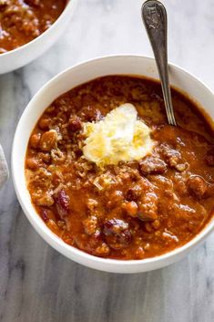 two bowls filled with chili and cheese on top of a marble counter next to a silver spoon