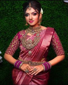 a woman in a pink and white sari with gold jewelry on her neck, standing next to a window