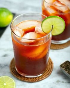 two glasses filled with ice and limes on top of a marble table next to sliced limes