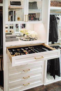 a white dresser with gold handles and drawers in a closet filled with clothes, jewelry and other items