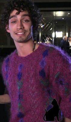 a man with curly hair wearing a purple sweater and smiling at the camera while standing next to a dog