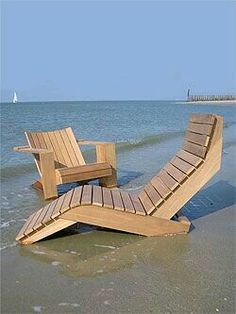 two wooden lounge chairs sitting on top of a sandy beach next to the ocean with a sailboat in the background