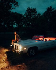 a woman sitting on the hood of a white car at night with trees in the background