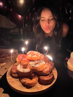 a woman blowing out candles on a cake with donuts in the foreground and balloons behind her