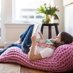 a woman laying on the floor reading a book while wearing a pink knitted blanket