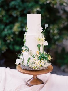 a three tiered white cake with flowers and greenery sits on a wooden stand