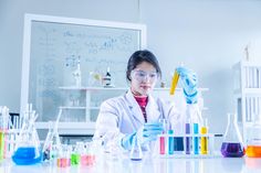 a woman in white lab coat and goggles working with flasks filled with colored liquid