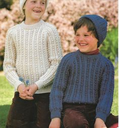 two young boys sitting next to each other in the grass wearing sweaters and hats