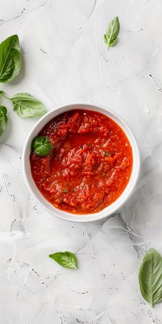 a bowl of tomato sauce with basil leaves around it on a marble table top, ready to be eaten