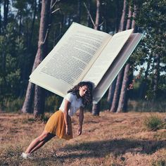 a woman is holding an open book over her head while standing in the grass with trees behind her