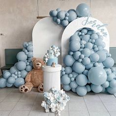 a teddy bear sitting in front of a balloon arch with blue balls on it and a cake